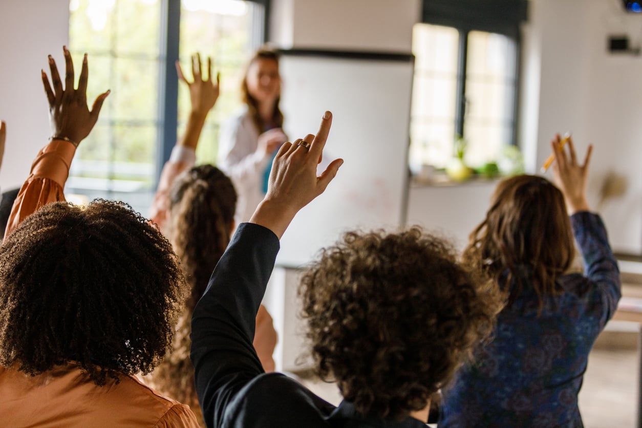 Diverse group of people raising hands in a classroom with a presenter.